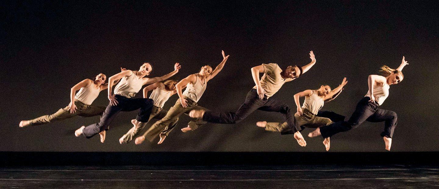 A group of dancers all leaping into the air, leaning right and extending their right arms and left legs.